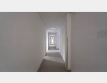 Langer, leerer Flur in einem Gebäude mit weißen Wänden, Teppichboden und mehreren geschlossenen Türen auf beiden Seiten. Am Ende des Flurs ist ein Fenster sichtbar, das für Tageslicht sorgt.