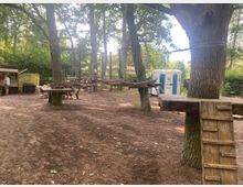 Ein Waldspielplatz mit Kletterelementen aus Holz, die zwischen Bäumen angebracht sind. Im Hintergrund stehen mobile Toiletten und ein kleiner Verkaufsstand, umgeben von dichtem Grün.