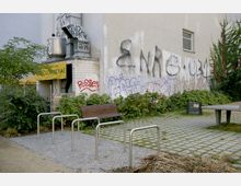 Ein kleiner städtischer Platz mit einem Holzparkplatz, einer Tischtennisplatte und Fahrradständern. Im Hintergrund eine Hauswand mit Graffiti und ein gelbes Schild mit der Aufschrift „Cucarachas“.