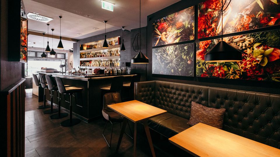 Modernes Bar- und Restaurantinterieur mit langem Tresen, Barhockern und beleuchteten Flaschenregalen. Rechts stehen Holztische vor einer gepolsterten Sitzbank, darüber hängen schwarze Pendelleuchten und großformatige Wandbilder mit Pflanzen- und Gewürzmotiven.