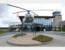 Gebäude des Flugplatzes "Schönhagen" mit einer verglasten Fassade und einem angebauten Kontrollturm aus grauem Beton. Im Vordergrund ist ein Hubschrauber auf einer Metallkonstruktion ausgestellt, umgeben von einem gepflasterten Weg und einem Kiesbeet mit niedriger Bepflanzung.