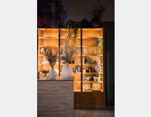 Holzregale mit warmer Beleuchtung, gefüllt mit dekorativen Gegenständen wie Keramikvasen, geflochtenen Körben und Pflanzen. Im Vordergrund steht ein Tresen mit weißen Vasen, die mit Trockenblumen arrangiert sind.