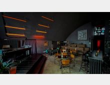 Innenraum einer modernen Bar mit dunkler Atmosphäre. Der Raum verfügt über eine Bühne, eine gewölbte Decke, orange LED-Lichtleisten an der Wand, Tische mit Holzstühlen und Bänke an der Rückwand; die Beleuchtung ist dezent mit einzelnen Kerzen.