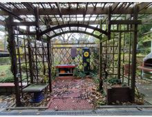 Holzpavillon in einem Garten, umgeben von Kletterpflanzen und Laub. Im Hintergrund befinden sich ein bemaltes Pfeilerobjekt mit Sonnenblume-Motiv, eine Rankhilfe und ein kleiner Fensterbereich mit Abstellfläche.