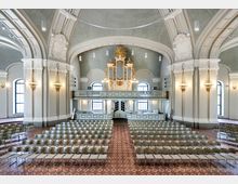 Innenraum einer historischen Kirche mit gewölbter Decke, verzierten Säulen und großen Rundbogenfenstern. Der Raum ist mit Stuhlreihen gefüllt, und eine barocke Orgel mit goldenen Ornamenten befindet sich auf einer Empore über dem Eingang.