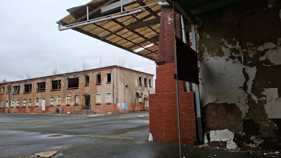 Verlassener Industriehof mit einem zweigeschossigen Backsteingebäude, dessen Fenster größtenteils zerstört oder vernagelt sind. Im Vordergrund sind eine bröckelnde Wand, ein roter Ziegelpfeiler und ein teilweise erhaltenes Vordach über einer großen Asphaltfläche zu sehen.