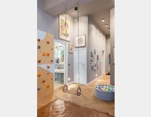 Ein heller Spielbereich mit einer Holz-Kletterwand links, einem Holz-Trapez, einem runden Bällebad rechts und einem Parkettboden. Im Hintergrund sind bunte Bilder an der Wand und ein angrenzendes Badezimmer mit Fliesen sichtbar.