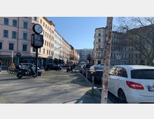 Straßenszene in einer Stadt mit gepflastertem Gehsteig und parkenden Autos, umgeben von mehrstöckigen Altbauten in verschiedenen Farben. Im Hintergrund ist das Schild eines Möbelgeschäfts ("Möbel Olfe") auf einem modernen Gebäude zu sehen, während vorne eine Uhr und Rollerparkplätze sichtbar sind.