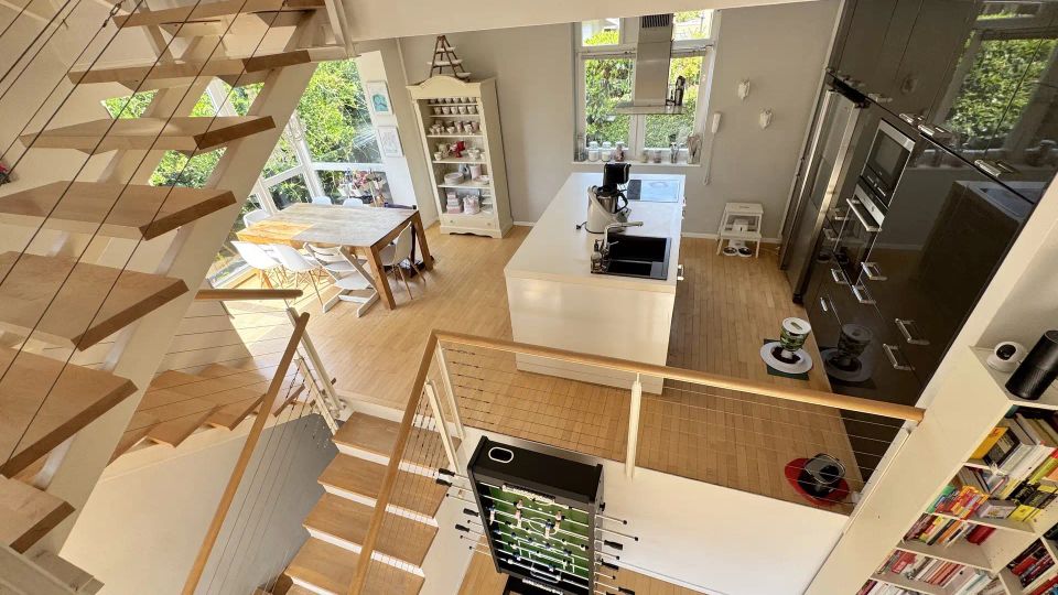Offener, moderner Wohn- und Küchenbereich über zwei Ebenen mit Holztreppe, Geländern aus Drahtseilen und hellen Holzböden. Große Fenster, eine zentrale Kücheninsel, ein Esstisch sowie Einbauschränke und Regale prägen den lichtdurchfluteten Raum.