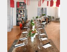 Ein gemütlicher Raum mit zwei langen Holztischen, die mit Weingläsern, Servietten, Besteck und Blumenarrangements dekoriert sind. Im Hintergrund befinden sich eine Bücherwand, ein Kunstwerk an der weißen Wand und verschiedenartige Stühle; rote Stoffbänder hängen von der Decke.