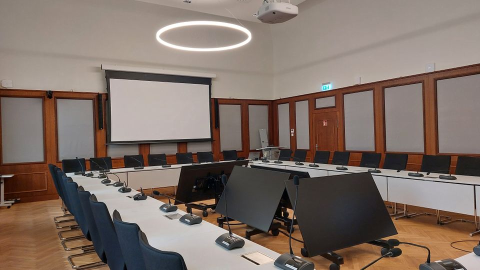 Moderner Konferenzraum mit langen, weißen Tischen in U-Form, ausgestattet mit Mikrofonen und Monitoren. Im Hintergrund befinden sich eine Projektionsfläche, Holzwandpaneele und eine Tür, die von schallisolierten Elementen umgeben ist; eine kreisförmige Leuchte hängt von der Decke.