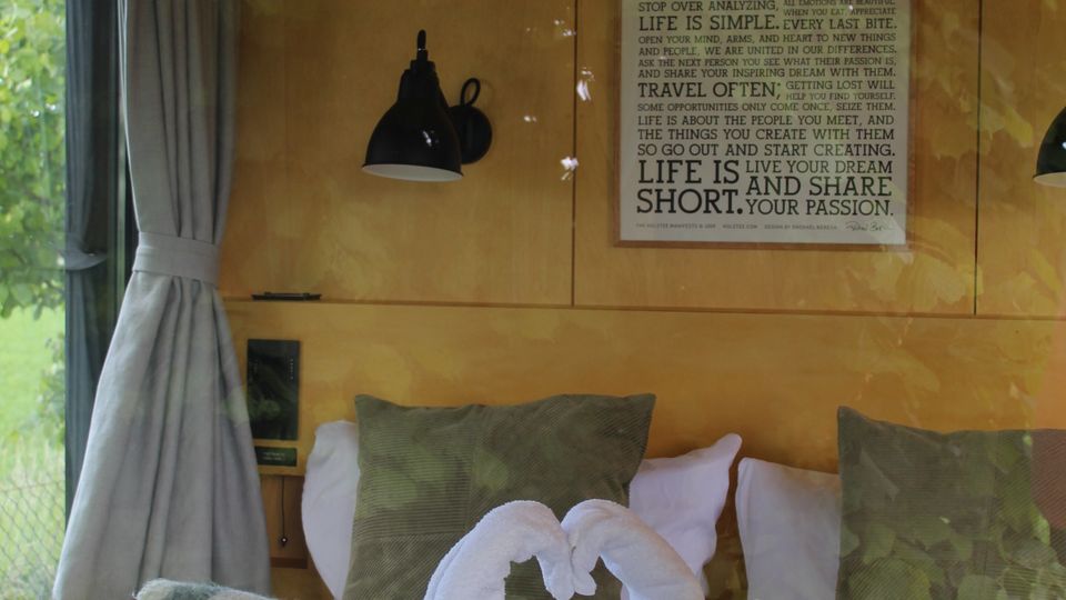 Innenansicht eines gemütlichen Schlafbereichs mit Doppelbett, gestreifter Decke und dekorativen Handtüchern in Herzform, flankiert von Kissen. An der Holzpaneelwand hängen zwei schwarze Wandlampen und ein typografisches Poster; links befindet sich ein großes Fenster mit grauen Vorhängen und Blick ins Grüne.