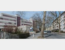 Straßenansicht eines mehrstöckigen Gebäudes mit grauer Fassade und roten Fenstern, das als medizinische Einrichtung ("Stiftstraße Klinikum") gekennzeichnet ist. Davor Gehwege, Autos und große, teilweise kahle Bäume; rechts angrenzende Wohngebäude.