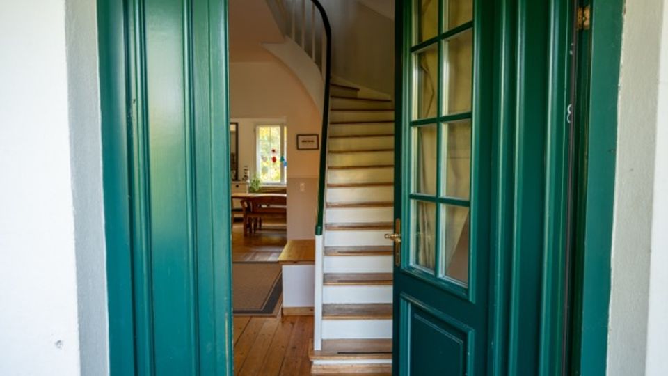 Blick durch eine geöffnete grüne Eingangstür in einen Hausflur mit Holzdielenboden und einer geschwungenen Treppe mit weißen Stufen und Holztrittkanten. Im Hintergrund ist ein heller Wohn- oder Essbereich mit Tisch sowie ein Fenster zu sehen.