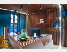 Moderner Innenraum mit einer Rezeption aus Holz, deren organisch geschwungene Lamellen in die Wand- und Deckenverkleidung übergehen, zusammen mit integrierter Beleuchtung. Im Hintergrund sind ein Fenster, dekorative Kunstwerke und ein Glasgefäß mit einer bepflanzten Miniaturlandschaft zu sehen.