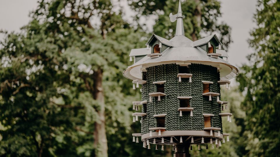 Zylindrisches Vogelhaus auf einem Pfahl mit schindelartiger Außenverkleidung und einem spitzen Metalldach mit Wetterfahne. Im Hintergrund sind unscharfe, dicht belaubte Bäume in einer parkähnlichen Umgebung zu sehen.