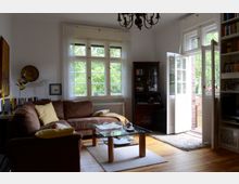 Wohnzimmer mit einer braunen Eckcouch, einem Glastisch auf einem Teppich und hölzernem Boden. Große Fenster und eine geöffnete Tür führen auf einen Balkon mit Blick ins Grüne, ergänzt durch Bücherregale und eine Hängelampe an der Decke.