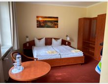 Hotelzimmer mit einem Doppelbett aus Holz, gelber Wandfarbe und rotem Teppichboden. Im Raum befinden sich ein Kleiderschrank, ein runder Holztisch mit einer Wasserflasche und zwei Gläsern sowie eine Fensterfront mit weißen Vorhängen.