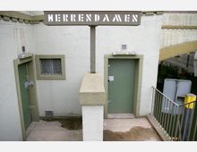 Öffentlicher Toiletteneingang mit separaten Türen für "Herren-WC" und "Damen-WC", beide in grün gehalten und in eine helle Betonwand eingelassen. Darüber befindet sich ein Schild mit der Aufschrift "HERREN DAMEN", daneben eine Treppe mit Geländer, die zu einem Wasserkanal führt.