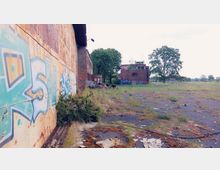 Verlassene Industrieanlage mit graffitibeschmierten Backstein- und Metallwänden, umgeben von einer unebenen, teils mit Gras und Moos bewachsenen Betonfläche. Im Hintergrund steht ein weiteres verfallenes Backsteingebäude, umgeben von Bäumen.