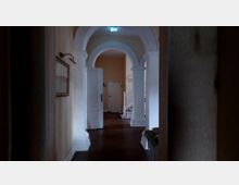 Flur in einem historischen Gebäude mit weißen Türrahmen, verzierten Bögen und einer geschwungenen Treppe auf der rechten Seite. Der Boden besteht aus dunklem Holz, und eine geöffnete Tür führt zu einem weiteren Raum im Hintergrund.