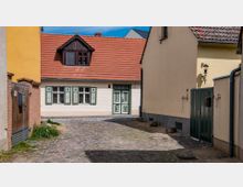 Eine gepflasterte Gasse mit Blick auf ein kleines, einstöckiges Haus mit hellem Putz, grünumlaufenden Fensterläden und einer auffälligen grünen Eingangstür. Das Haus hat ein rotes Satteldach mit einem mittig platzierten Dachfenster, umgeben von gelben und beigen Fassaden anderer Gebäude.