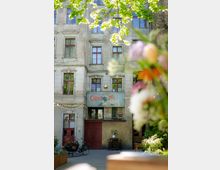 Alt-Text: Ein Altbau mit einer Fassade aus hellem, gealtertem Putz und roten Fensterrahmen. Über dem Eingang befindet sich ein Schild mit der Aufschrift "Clärchens Ballhaus". Im Vordergrund sind Gebüsch und bunte Blumen unscharf zu sehen, und der Hof ist mit Tischen, Stühlen und einem Lastenfahrrad ausgestattet.