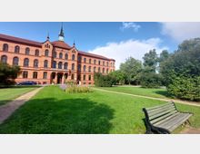 Backstein-Gebäude im neogotischen Stil mit Turm und Uhr, umgeben von einer gepflegten Grünanlage mit Wegen, Bäumen und Büschen; im Vordergrund eine leere Holzbank auf einer Wiese.