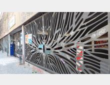 Metallzaun mit abstraktem, organischem Muster vor einem Parkhaus, das teilweise sichtbar ist. Die Zaunelemente sind mit Graffiti und Aufklebern versehen; im Hintergrund sind ein gepflasterter Gehweg und ein Gebäude mit Klinkerfassade erkennbar.