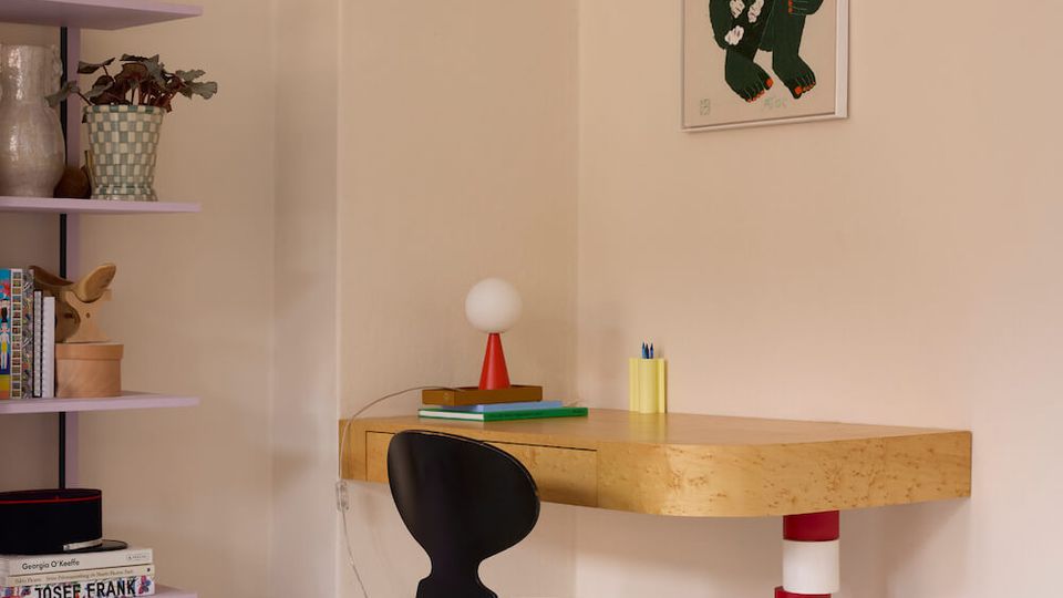 Minimalist home office corner with a light wood floating desk supported by a red-and-white striped column, paired with a black chair on a white plank floor. A lavender shelving unit sits to the left, and a framed graphic artwork hangs on the beige wall above the desk.