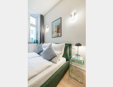 Modernes Schlafzimmer mit grün gepolstertem Bett, weißer Bettwäsche und mehreren Kissen, daneben ein gläserner Nachttisch mit schwarzer Lampe. Helle Wände mit zwei Wandleuchten und einem „Berlin“-Poster, großes Fenster mit grauem Vorhang sowie Holzboden.