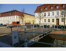 Straßenszene mit historischen Wohnhäusern in heller Putzfassade und roten Ziegeldächern, davor geparkte Autos und städtische Fahrzeuge. Im Vordergrund eine kleine Metallbrücke über einen Wasserlauf mit Geländern und Maschendrahtzaun.