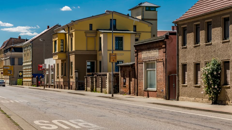 Straßenszene in einer Kleinstadt mit zweispuriger Fahrbahn und Gehweg, flankiert von mehrgeschossigen Wohn- und Geschäftshäusern mit Putz- und Backsteinfassaden sowie Satteldächern. Mehrere Verkehrsschilder und eine Bushaltestelle stehen am Straßenrand, der Himmel ist blau mit vereinzelten Wolken.