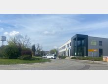 Moderne Bürogebäudefassade mit schwarzem Eingangsbereich und großen Fenstern rechts im Bild; im Vordergrund eine Straße mit Kreisverkehr, grünen Rasenflächen und einer Standuhr; im Hintergrund Bäume und einige geparkte Autos.