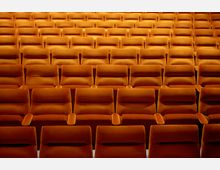 Reihen von gepolsterten, orangefarbenen Sitzen in einem Theater- oder Kinosaal, mit hoher Symmetrie und regelmäßigem Abstand.