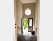 Treppenhaus mit grauen Wänden, das zu einer offenen doppelflügeligen Haustür führt. Über der Tür befindet sich ein rundes Fenster, das Tageslicht einlässt, und hinter der Tür ist ein kleiner gepflasterter Hof mit grüner Vegetation sichtbar.