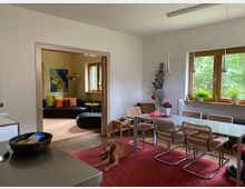 Wohnbereich mit Esszimmer und angrenzendem Wohnzimmer. Im Vordergrund ein Esstisch mit Stühlen auf einem roten Teppich, daneben ein ruhender Hund; im Hintergrund ein Wohnzimmer mit Sofa, Dekoration und einem großen Fenster mit Blick ins Grüne.
