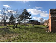 Ländliches Grundstück mit grüner Wiese, kahlen Bäumen und vereinzelten Kiefern. Rechts im Vordergrund sind rote Backsteingebäude mit Ziegeldächern, und im Hintergrund steht ein weiteres Backsteingebäude. Der Himmel ist klar mit einigen Wolken.