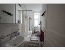 Helles Badezimmer mit weißer Badkeramik, einer Badewanne mit Duschvorhang, einem Waschbecken und einer Toilette unter einem Fenster mit Tageslicht. Ein Handtuchheizkörper an der Wand und Teppiche auf dem Boden ergänzen die Einrichtung.