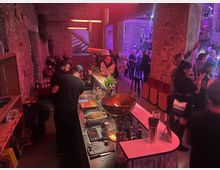 Industrie-inspirierte Bar in einer Halle aus rohem Mauerwerk mit roten und violetten Lichtakzenten; im Vordergrund eine Theke mit Zutaten und Getränken, im Hintergrund eine Menschenmenge bei einer Veranstaltung.
