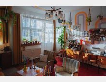 Wohnzimmer mit Retro-Einrichtung, roten Teppichboden und großen Fenstern mit halbtransparenten Vorhängen. Pflanzen und Dekorationen füllen den Raum, mit Wandregalen, einem Schreibtisch, einer Nähmaschine und einem Couchtisch mit Kerzen als zentrale Elemente.