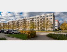 Wohnblock einer Mehrfamilienhaussiedlung mit fünf Etagen, umgeben von Bäumen mit herbstlichem Laub. Im Vordergrund befinden sich ein Parkplatz, gepflasterte Wege und gepflegte Grünflächen.