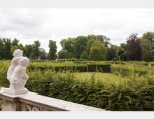 Steinbalustrade mit einer weißen Skulptur zweier spielender Kinder im Vordergrund, dahinter gepflegte Hecken und eine weitläufige Parklandschaft mit Grünflächen und hohen Bäumen. Der Himmel ist bewölkt.