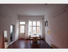 Ein einfacher, weiß gestrichener Raum mit einem rötlich-braunen Boden, einem großen Fenster mit durchsichtigem Vorhang sowie einer Glastür, durch die Tageslicht einfällt. In der Mitte steht ein Holztisch mit drei Hockern, an den Wänden befinden sich ein Spiegel, ein kleines Wandbrett und ein Bild.