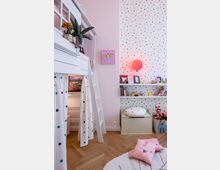 Kinderzimmer mit pastellrosa Wänden und einer weißen Hochbett-Konstruktion mit Leiter. Die Wand ist teils mit bunten Punkten verziert, Regale mit Spielsachen und Büchern schmücken den Raum, während der Boden aus hellem Holzparkett besteht.