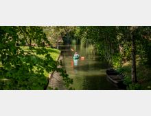 Eine Person paddelt in einem grünen Kajak durch einen schmalen, von Bäumen und Gras gesäumten Fluss. Am Ufer liegt ein altes Holzboot, und die Szene ist von dichtem Blattwerk umgeben.