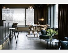 Moderner Innenraum eines Cafés oder Lounge-Bereichs mit großen Fenstern, einem dunklen Tresen samt Barhockern, einem Holztisch mit passenden Stühlen und einer Sitzecke mit Sofas. Eine Vase mit Blumen steht auf einem Beistelltisch und der Raum ist mit Teppichen und schlichten Hängelampen dekoriert.
