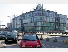 Ein modernes Einkaufszentrum mit einer Glasfassade und mehreren Logos, darunter "Saturn", an der Vorderseite. Im Vordergrund eine Straße mit parkenden und fahrenden Autos sowie Absperrungen entlang der Fahrbahn.