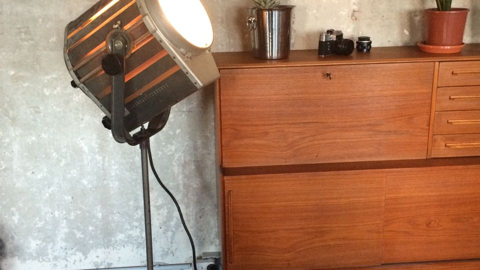 Innenraum mit rauer Betonwand und einem hölzernen Sideboard, auf dem kleine Pflanzen und Kameraobjektive stehen. Daneben steht ein großer, industrieller Scheinwerfer auf einem Stativ mit sichtbarem Kabel.