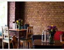 Gemütlicher Innenraum eines Cafés mit Sichtbacksteinwand, einfachen Holztischen und -stühlen. Auf den Tischen stehen Gläser mit bunten Blumenarrangements und Kerzen; ein Fenster links lässt Tageslicht herein.